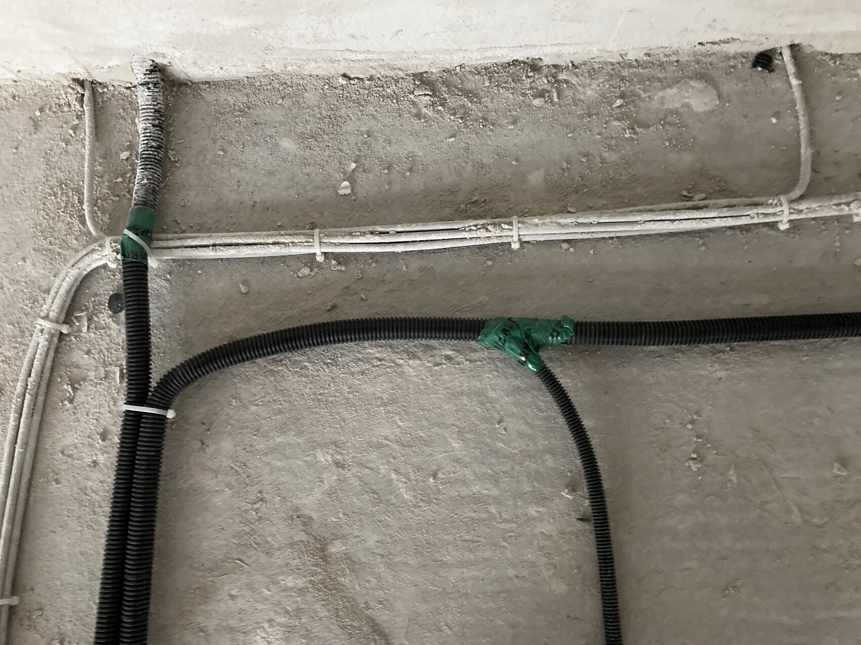 Verlegte schwarze Flexleitungen und weiße Kabelrohre an staubiger Betonwand.