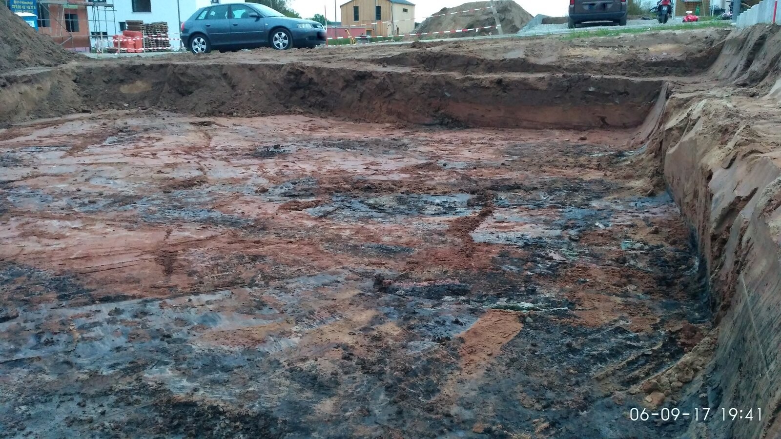 Grube auf Baustelle mit unterschiedlichen Erdschichten, Bauzubehör und parkenden Autos im Hintergrund