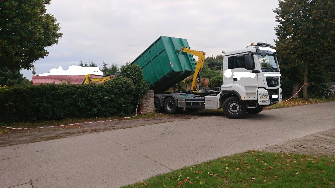 Weißer LKW mit grünem Container wird von gelbem Kran auf Anhänger gehoben.