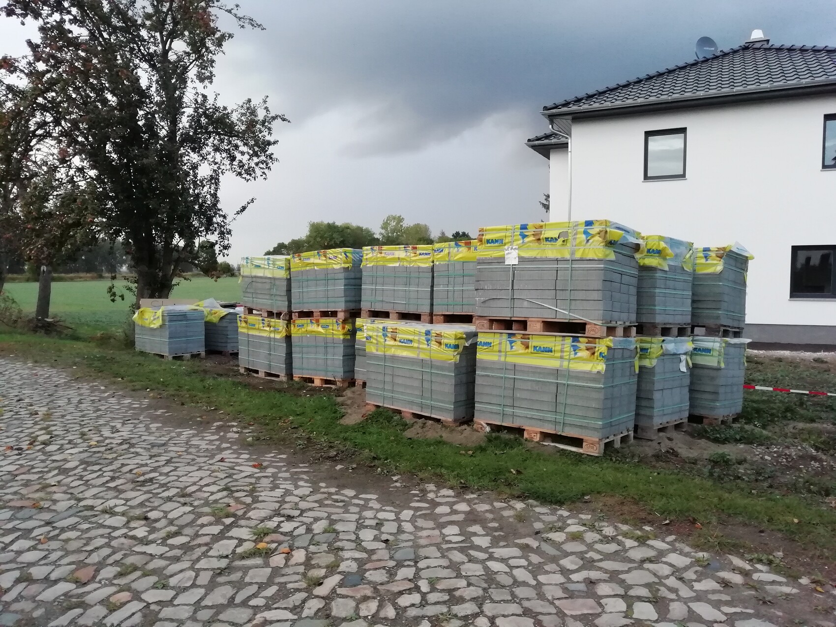 Mehrere Paletten Beton-Dachziegel am Rand eines weißen Hauses auf Kopfsteinpflasterweg.