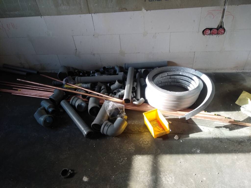 Haufen Rohre und Fittings aus PVC und Kupfer auf Betonboden, daneben eine große silberne Spule.