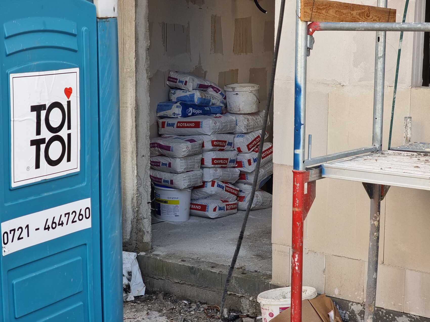 Bauplatz: blaue Toilettenkabine links, hinter Eingang gestapelte Rotband-Putzsäcke und Gerüst rechts.