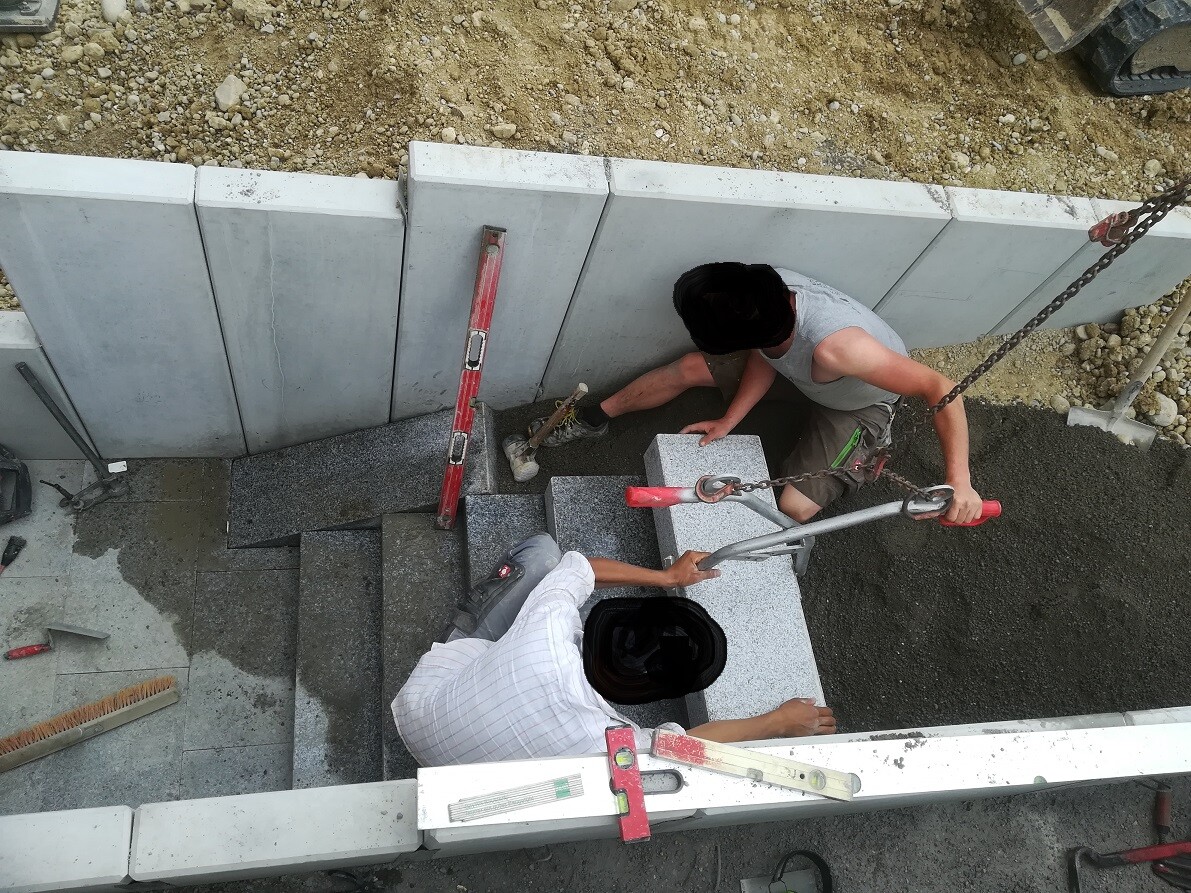 Zwei Bauarbeiter installieren Betonstufen an einer Treppe, mit Kranhaken und Werkzeugen.