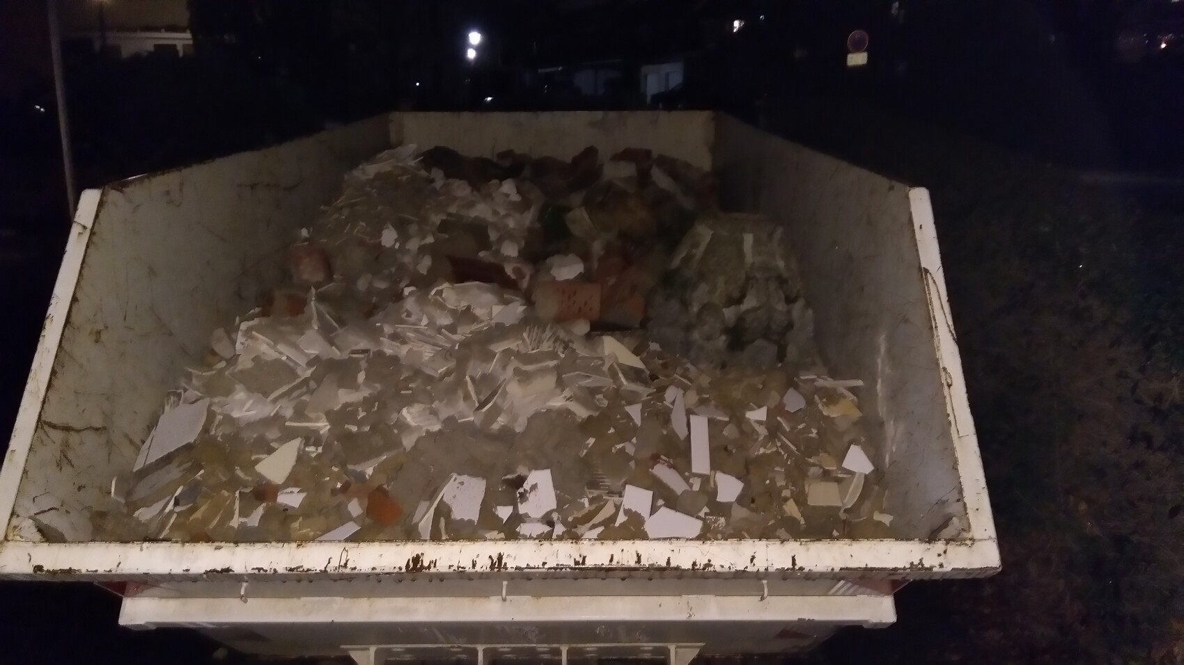 Nachtaufnahme eines vollen Abfallcontainers mit Bauschutt, Fliesen und Papier.