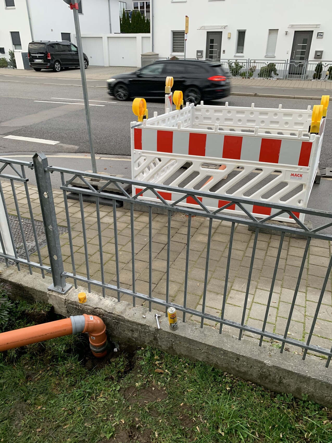 Baustellenabsperrung am Gehweg, weiße Barriere mit roten Streifen, orange Rohr, Auto fährt vorbei.