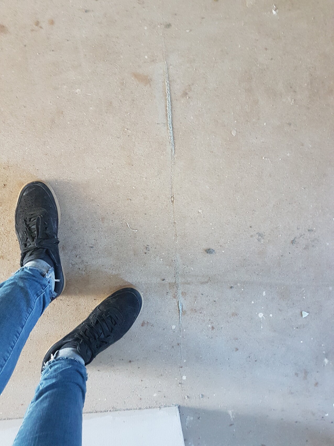 Obere Perspektive: Person in Jeans und schwarzen Sneakern steht auf rauem Betonboden mit Riss.
