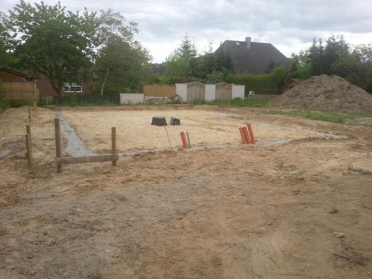 Baustelle mit Graben, Holzpfosten, orange Markierungen, Sandboden, Gartenhäuser im Hintergrund.