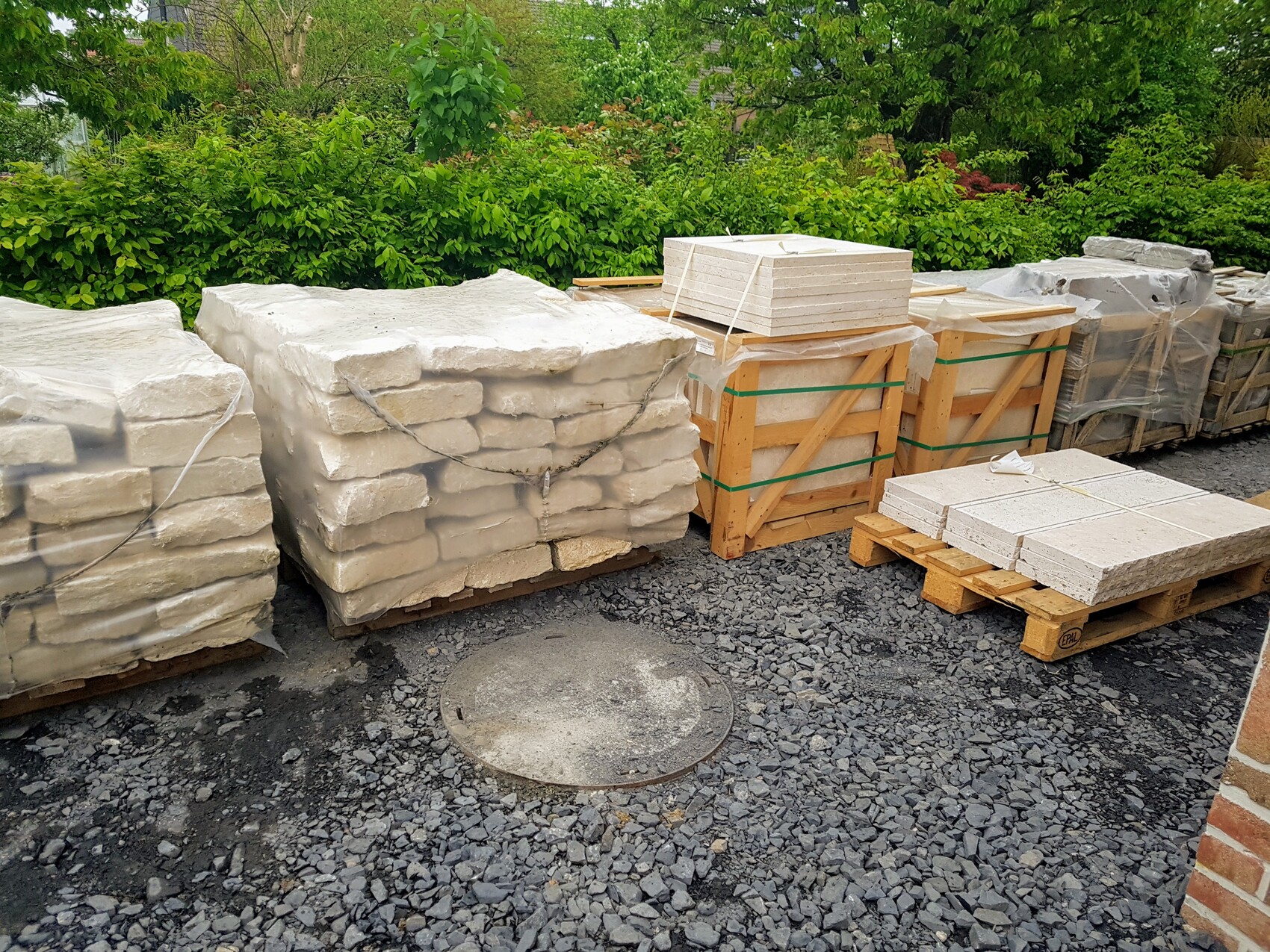 Große Baustoffstapel aus Steinplatten und Blöcken auf Paletten vor grüner Hecke.
