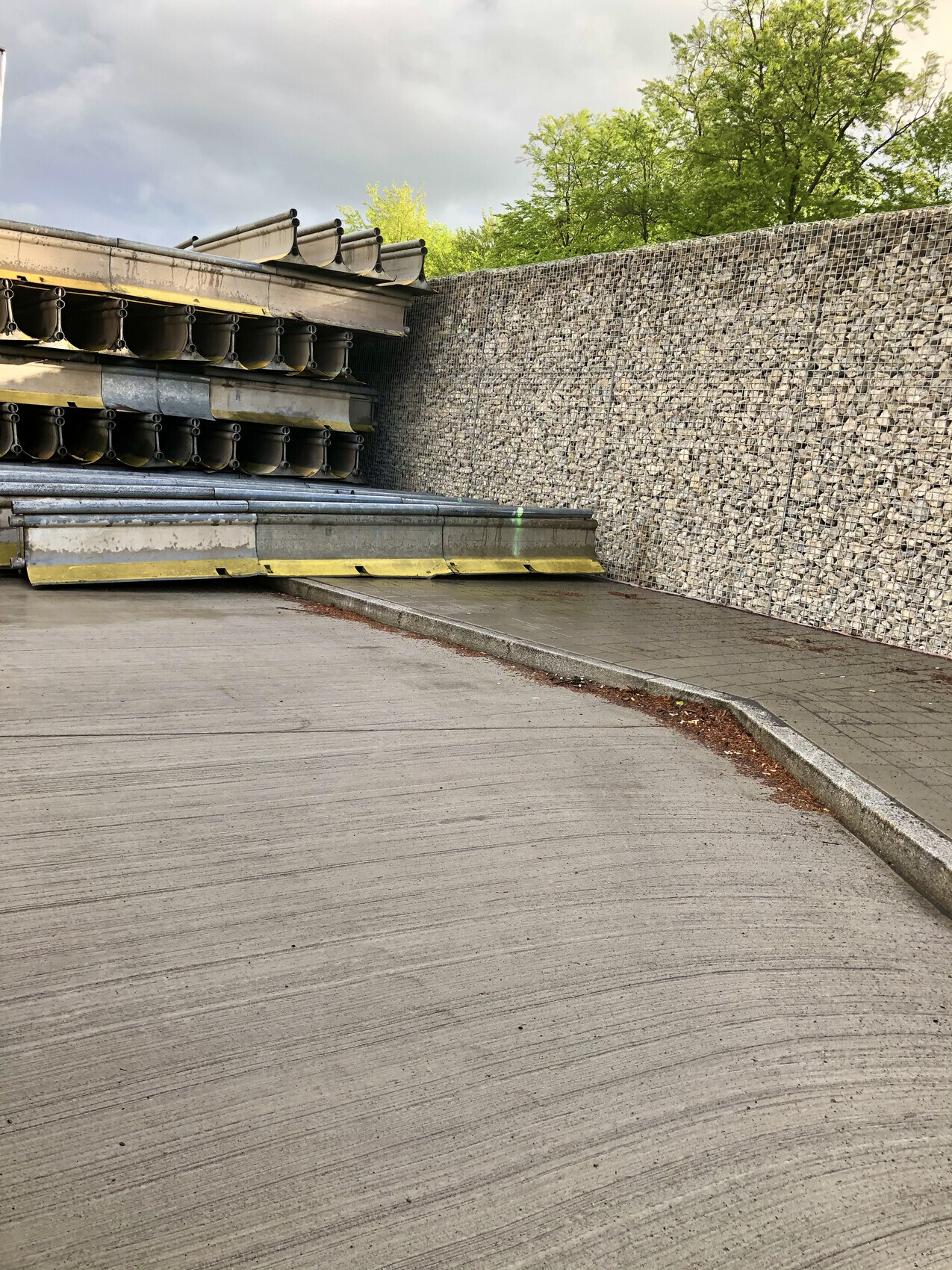 Gestapelte Metallrohre/Profile neben Steinmauer; Betonfläche vorne, grüne Bäume dahinter.