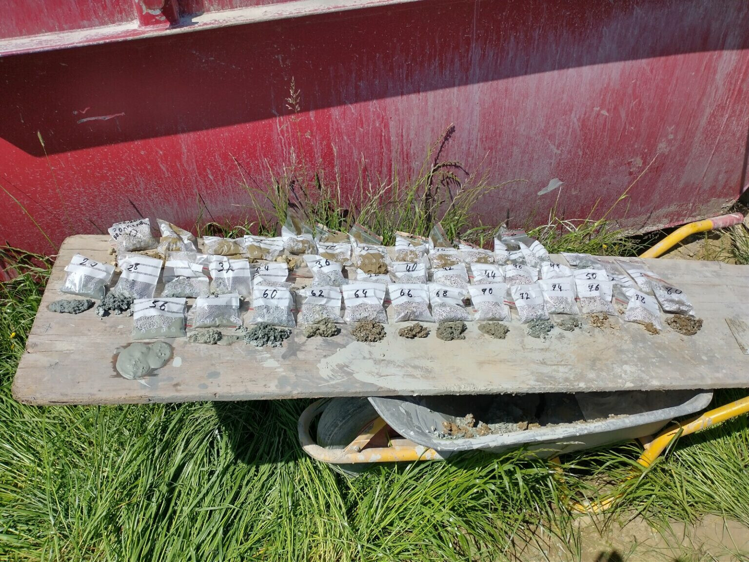 Mehrere nummerierte Probenbeutel mit grauen Beton-/Gestein-Proben auf Holztisch vor roter Wand.