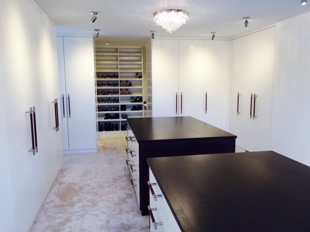 Heller begehbarer Kleiderschrank mit weißen Schränken, Schuhregale im Hintergrund, dunkle Inseln in der Mitte.