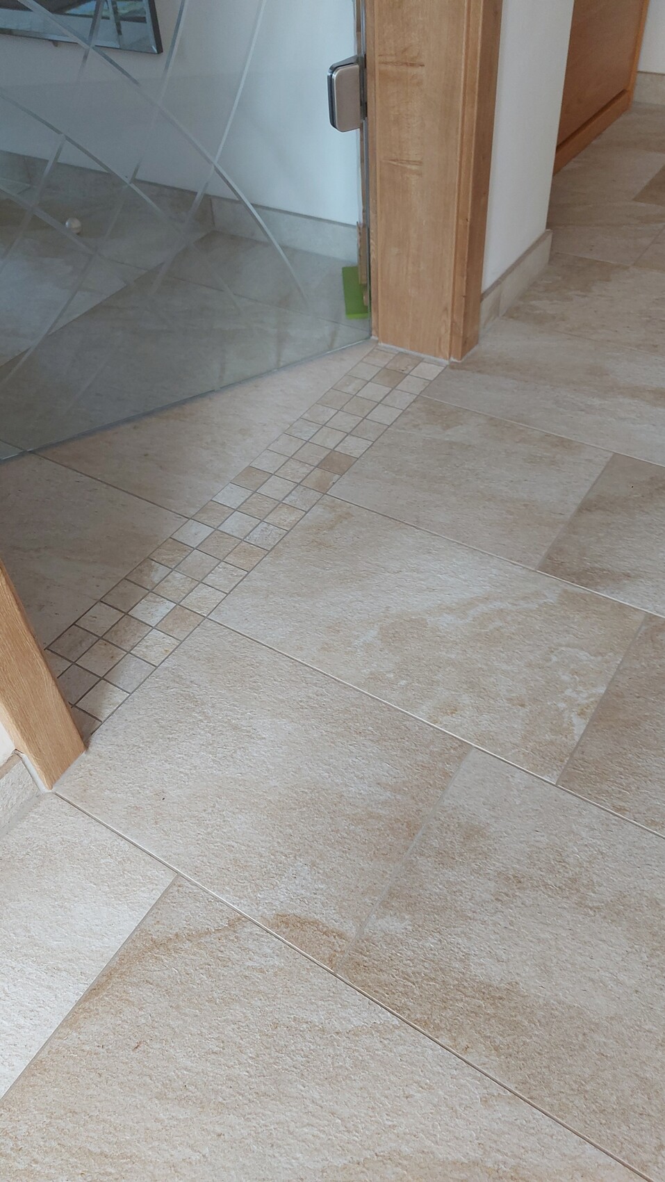 Beiger Fliesenboden mit schmaler Mosaikleiste am Türanschlag; Glastür und Holztürrahmen.