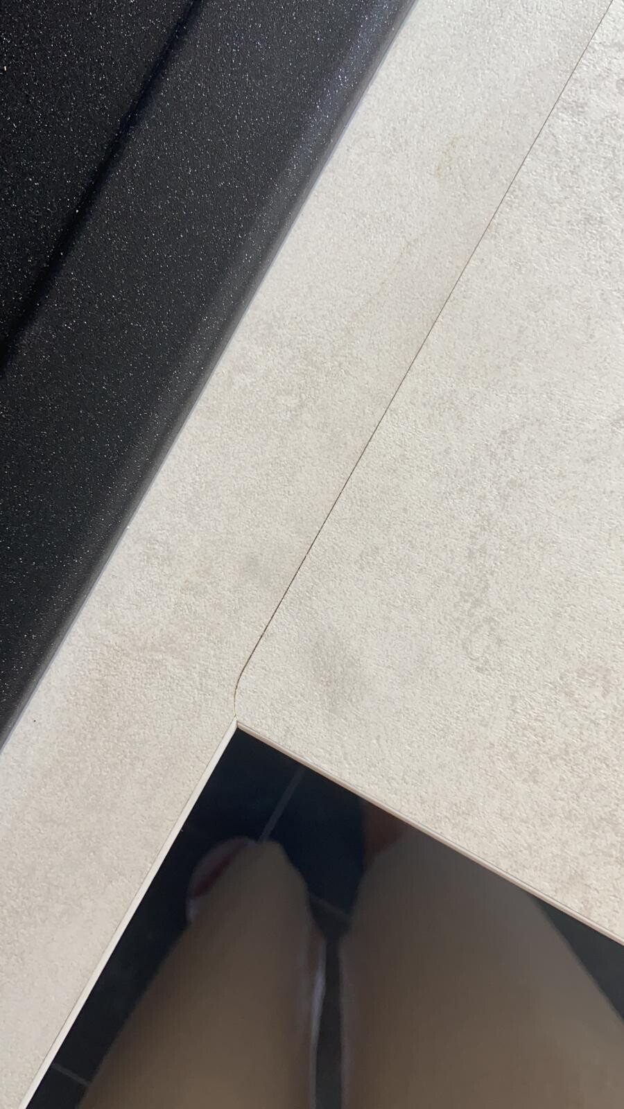 Nahaufnahme beiger Bodenfliesen neben dunkler, glänzender Fläche; diagonale Fuge mit Riss.