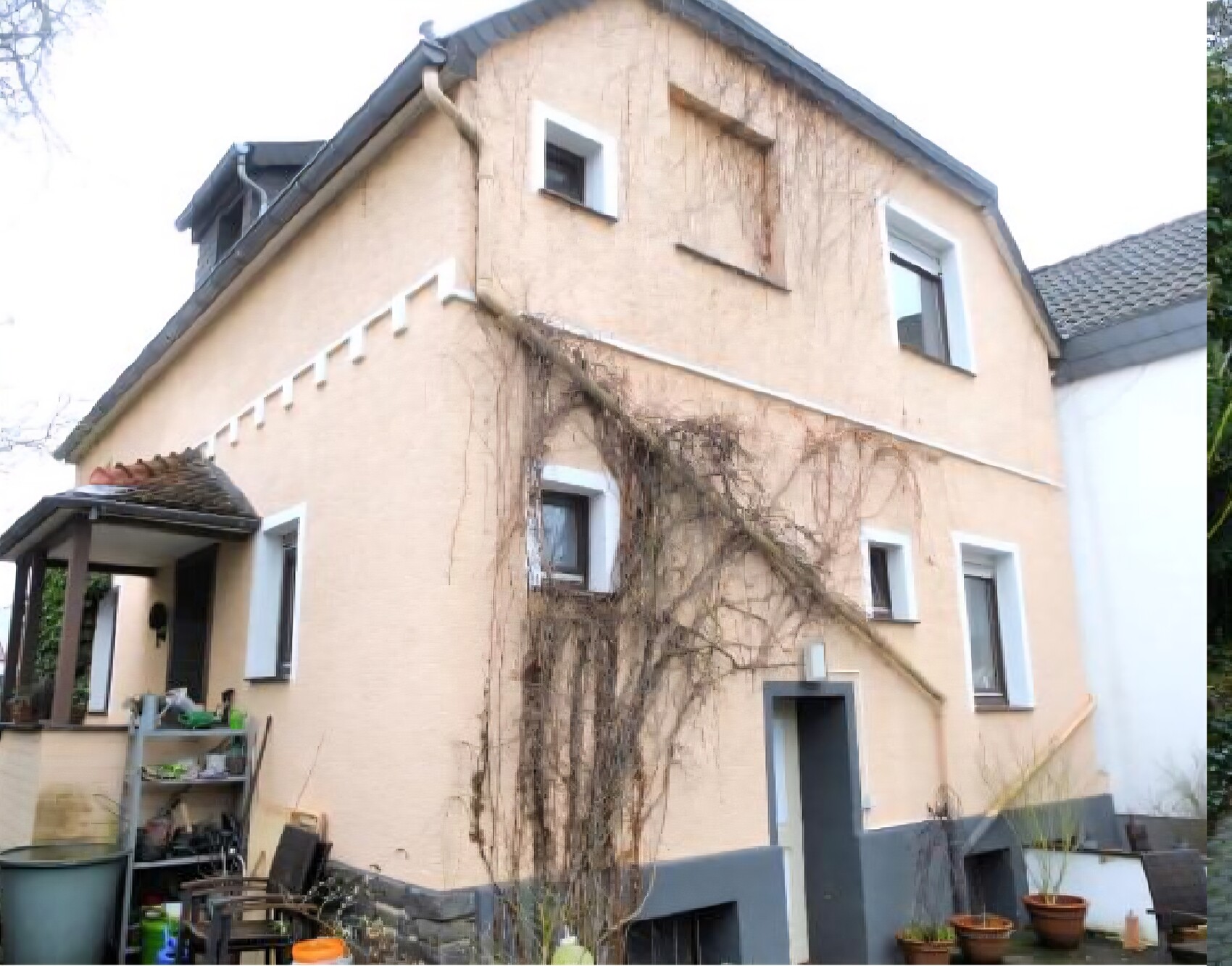 Beiges zweistöckiges Haus mit rankenden Pflanzen an der Wand und Gartengeräten im Vorgarten.