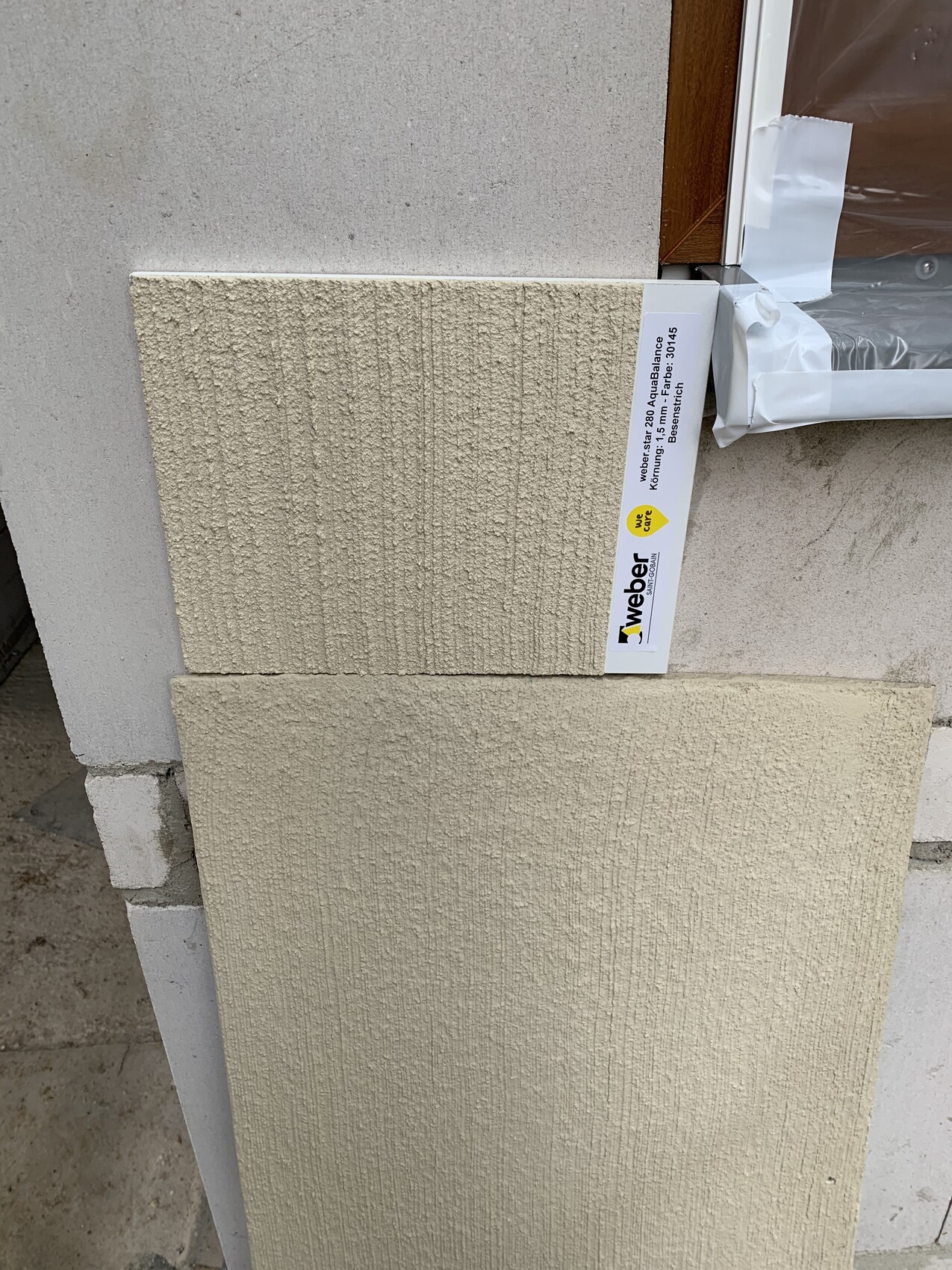 Zwei beige-texturierte Fassadenplatten lehnen an einer Außenwand; daneben Fensterrahmen mit Folie.
