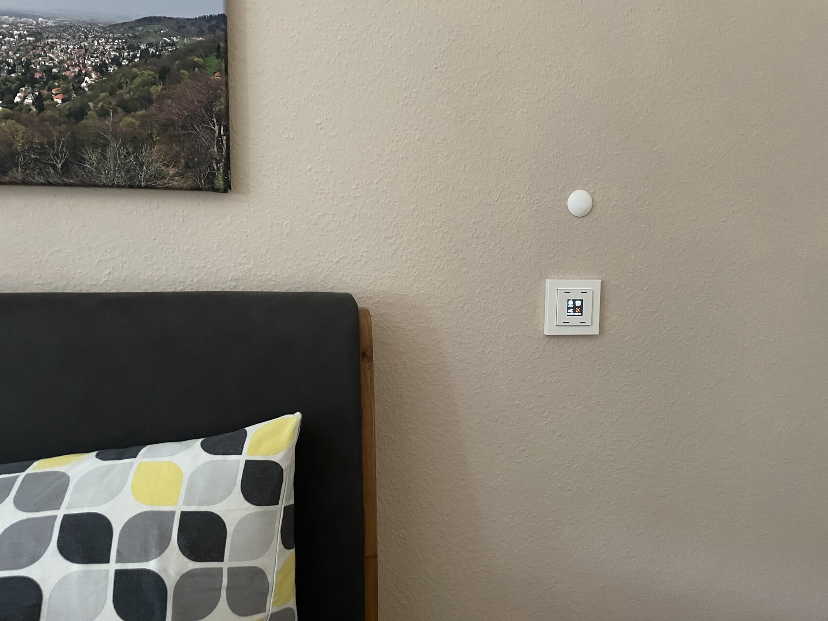 Schlafzimmerwand mit Bettkopfteil, geometrischem Kissen, Wandbild links, runder Sensor und Schalter.