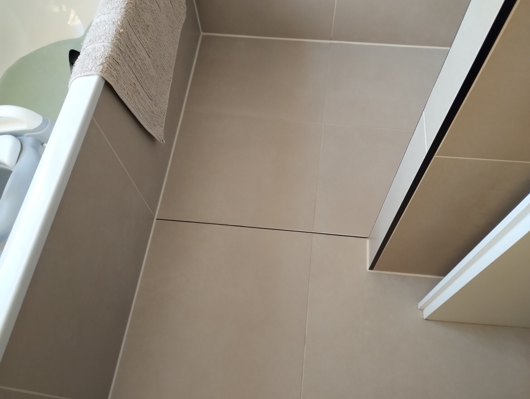 Beiges Badezimmer mit großen Bodenfliesen, Wannenrand und Duschtür.