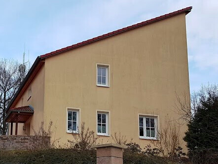 Außenansicht eines beigen Hauses mit rotem Ziegeldach, Fenstern und Sträuchern.