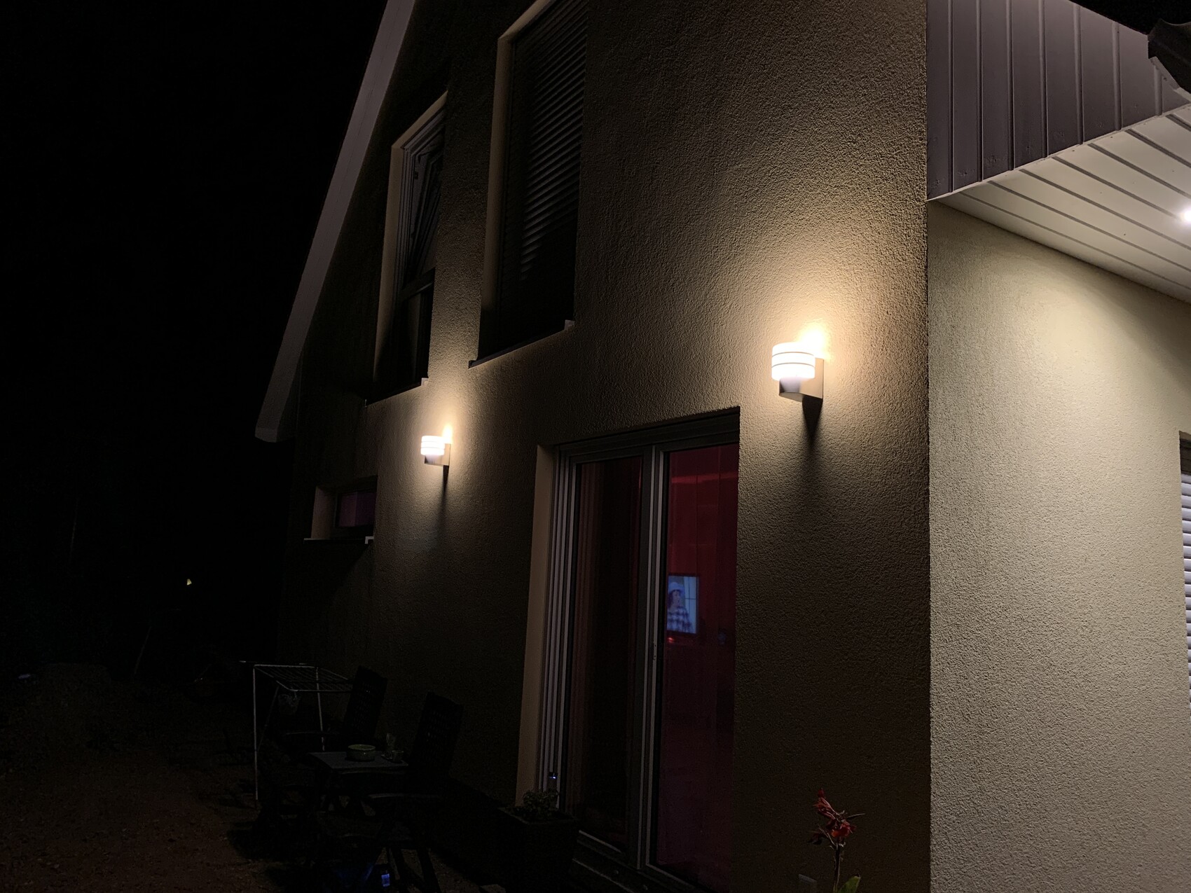 Außenansicht eines Hauses in der Nacht; warmes Licht aus Wandlampen, Terrasse mit Tisch und Stühlen