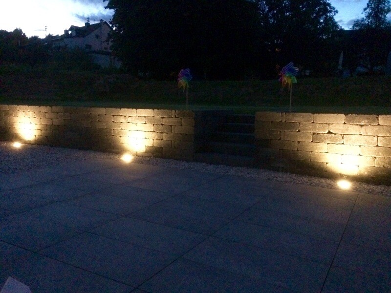 Abendliche Terrasse mit beleuchteter Steinmauer, Stufen und gepflastertem Vorplatz