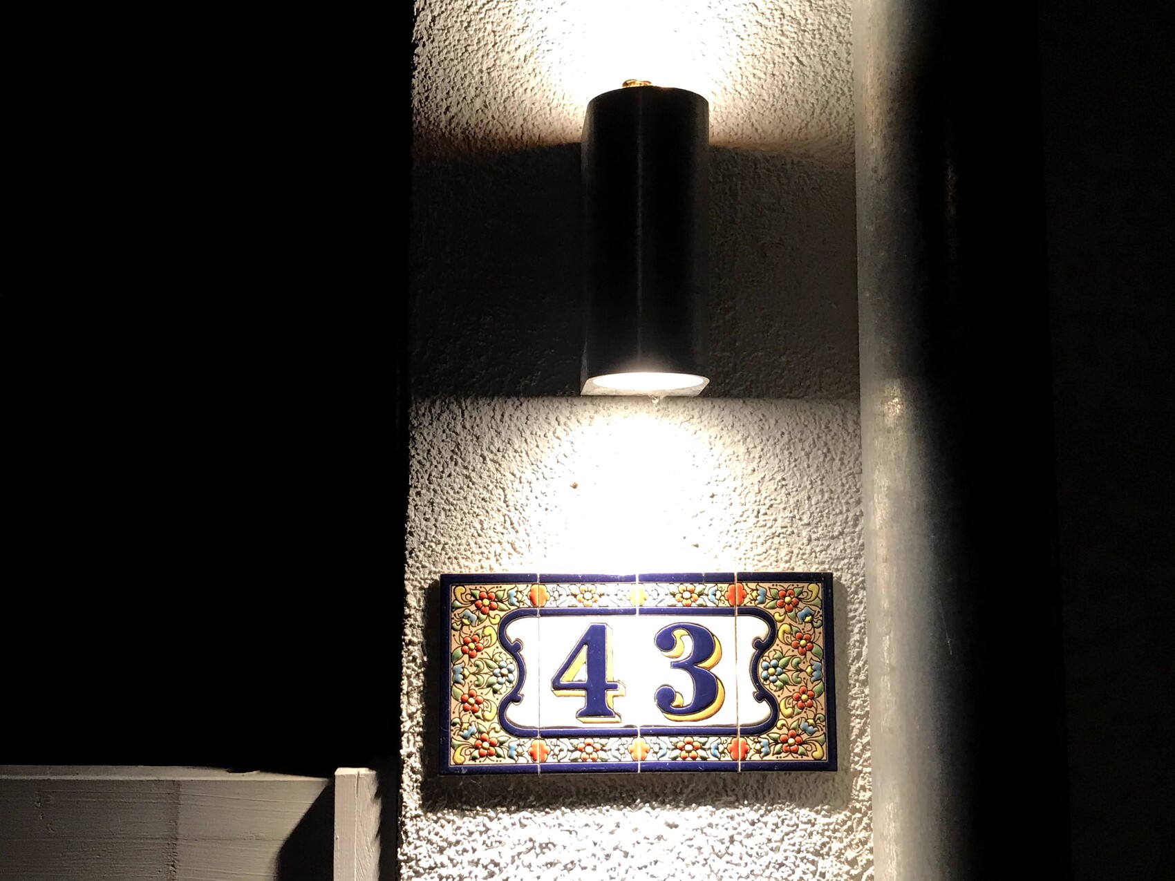 Keramische Fliesen-Hausnummer 43 mit buntem Blumenrand an rauer Wand, beleuchtet von einer Außenlampe.