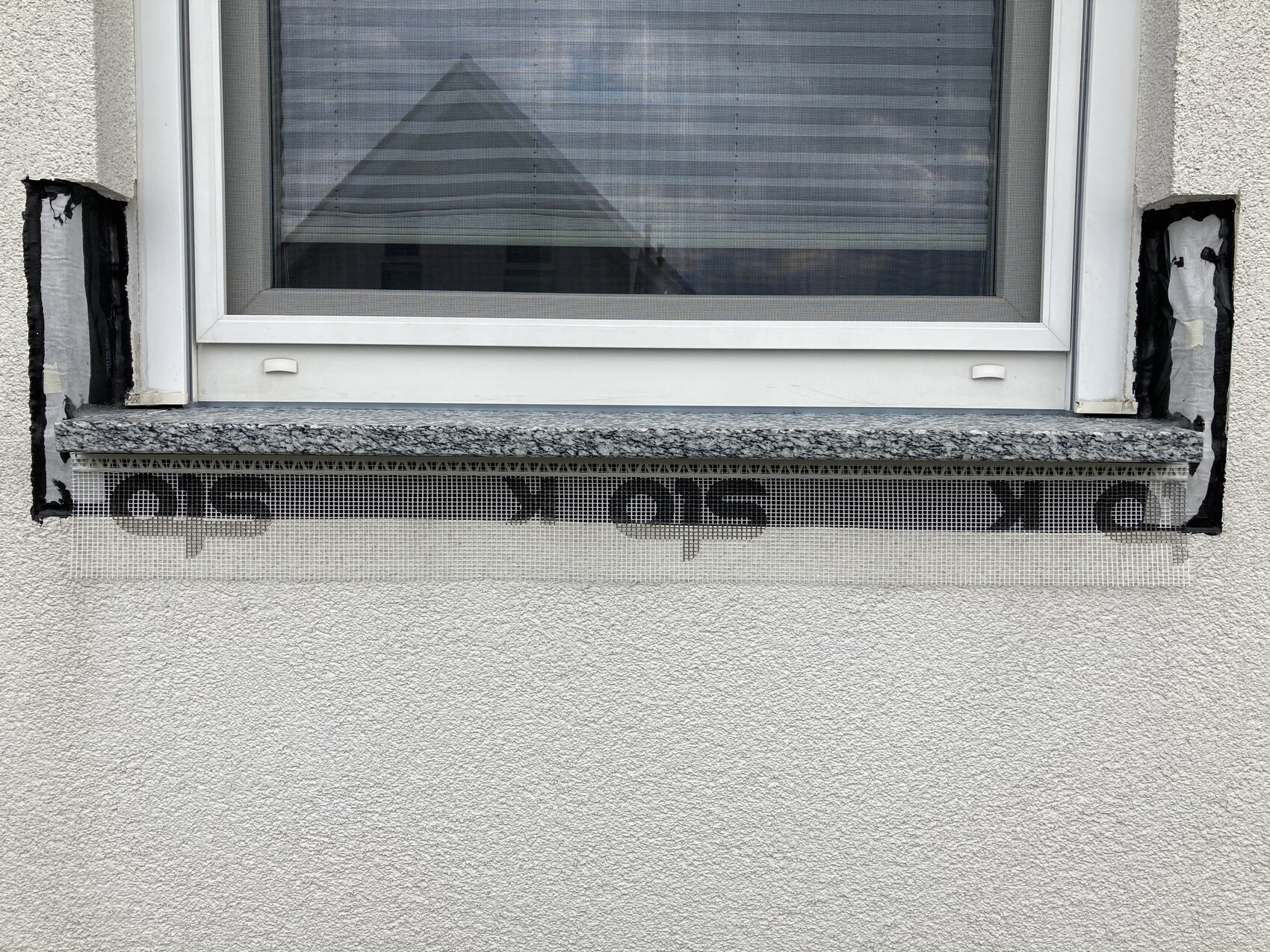 Fenster in heller Fassade; darunter eine Steinbank und ein Metallnetz mit Buchstaben.