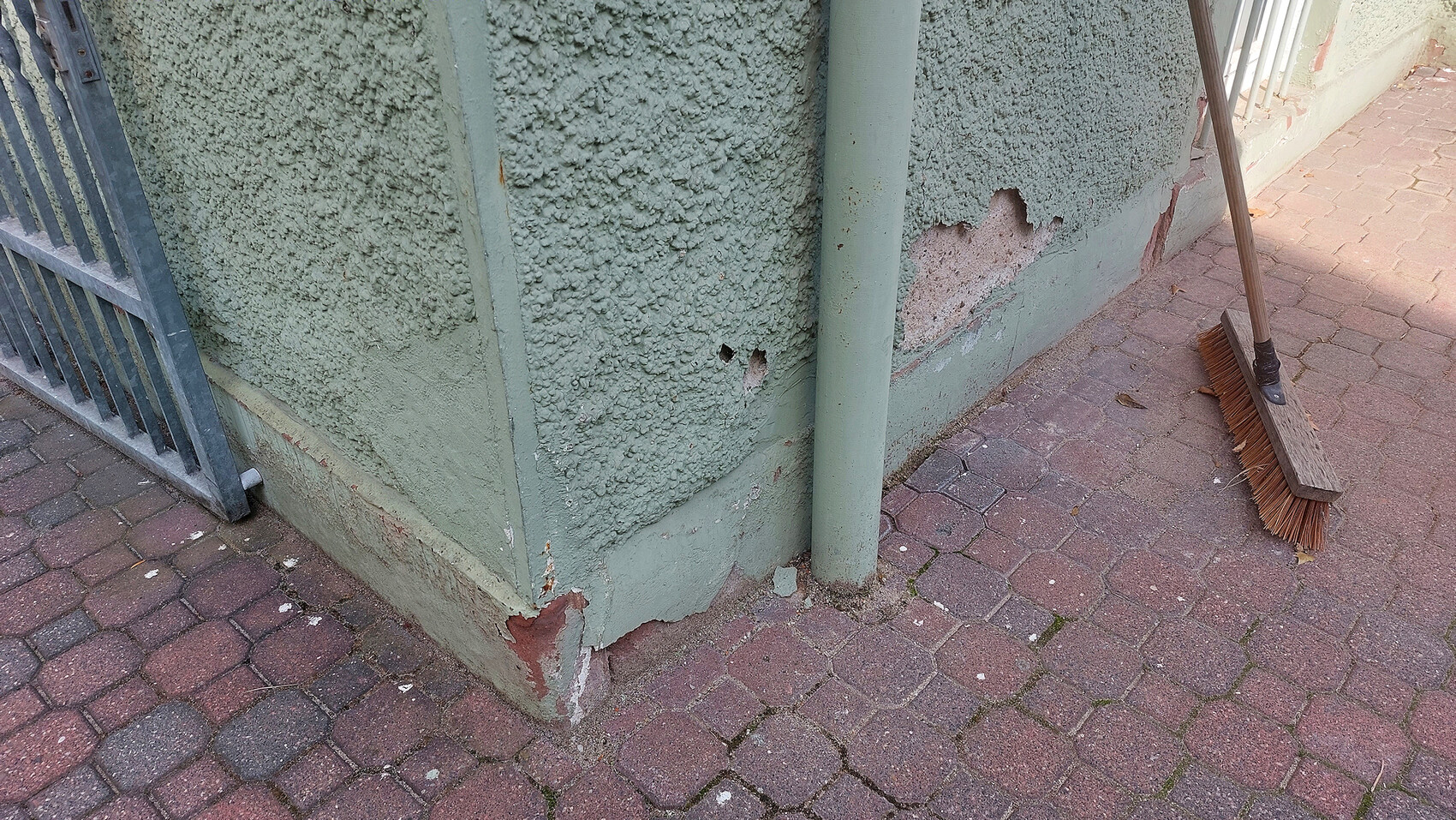 Außenwand mit rauer Putzstruktur, grüne Farbe blättert ab, Löcher, Abplatzungen; Besen am Boden.