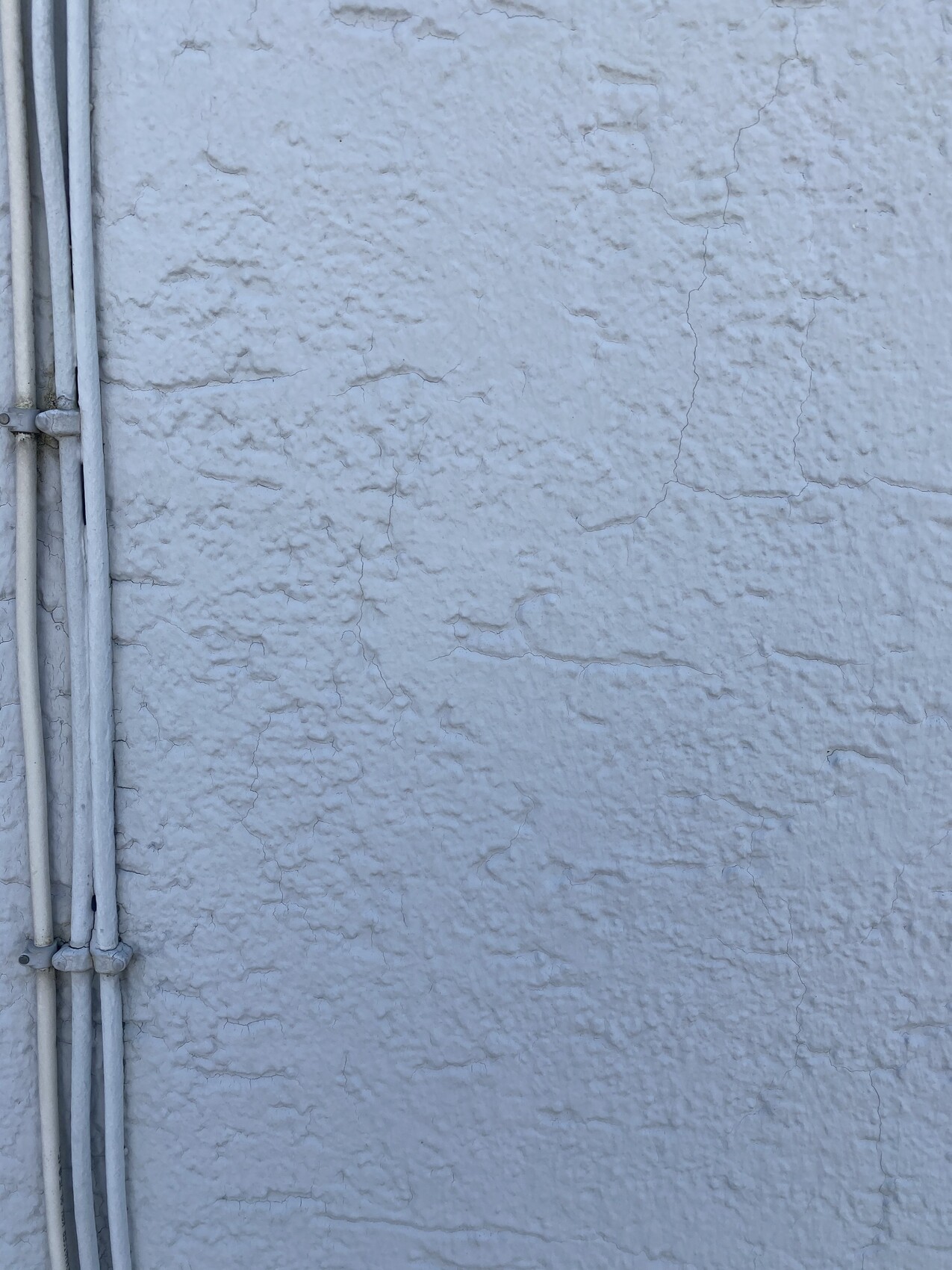 Weiße raue Verputzwand mit Rissen; links sichtbare Leitungsrohre.