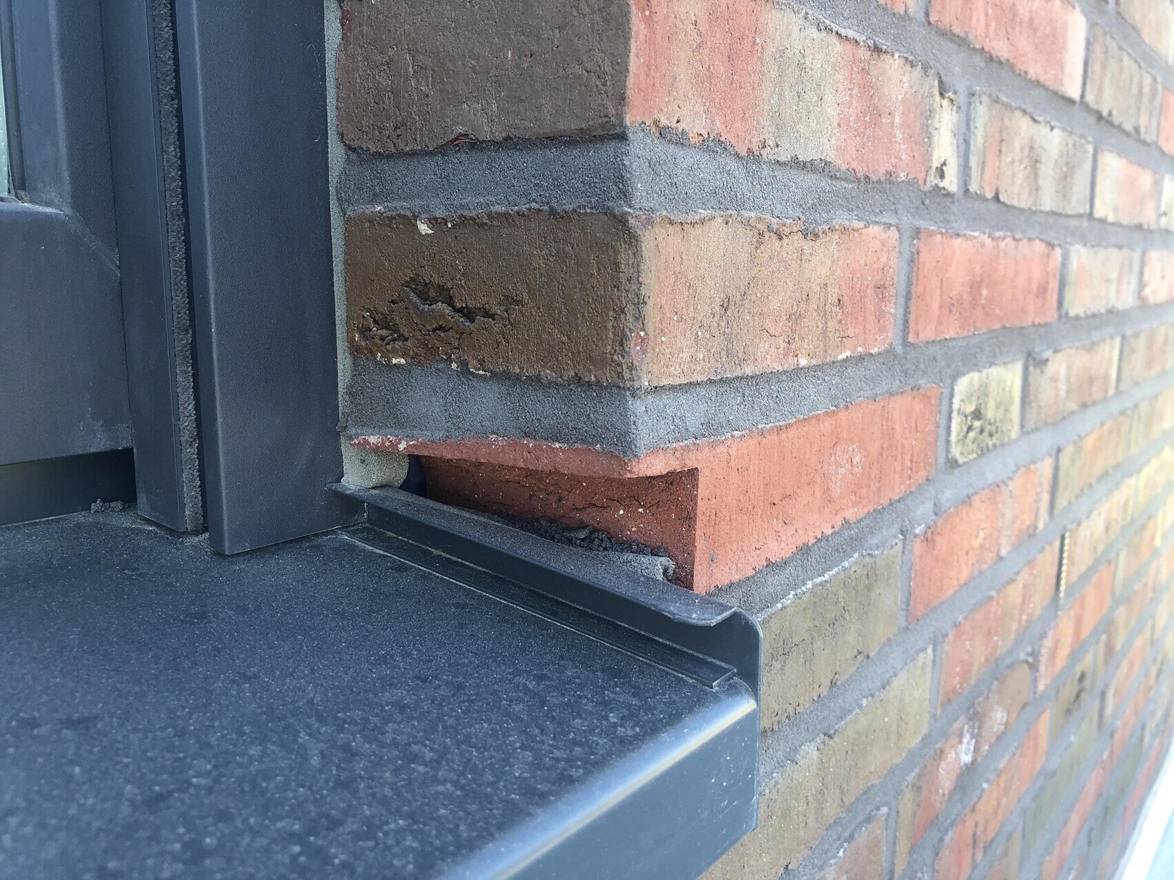 Nahaufnahme roter Backsteinwand mit beschädigtem Ziegel neben dunkler Fensterbank und Metallrahmen.