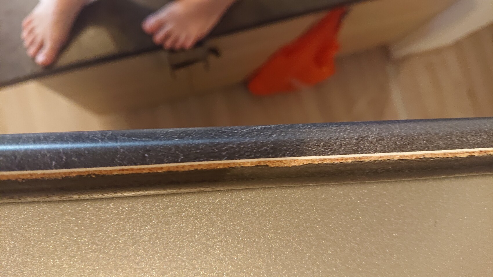 Nahaufnahme eines abgenutzten Möbelrands mit Kante; barfuß sichtbare Füße im Hintergrund.