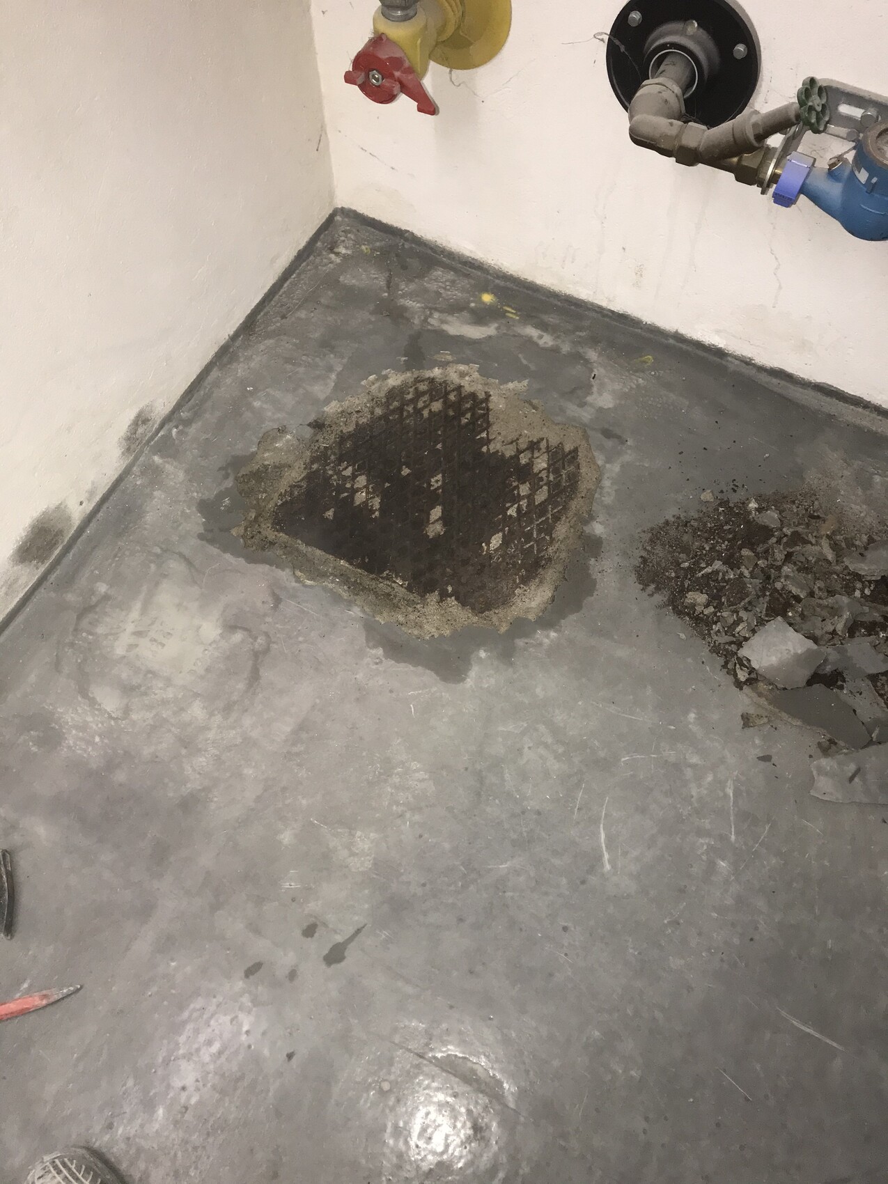 Boden mit Loch, freigelegtes Bewehrungsstahlgitter in Beton, Rohrleitungen an der Wand.