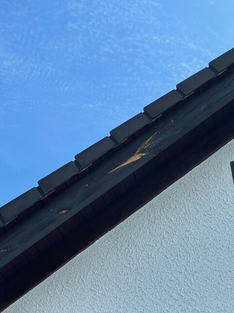 Nahaufnahme einer Dachkante mit dunklen Ziegeln über einer weißen Wand, blauer Himmel