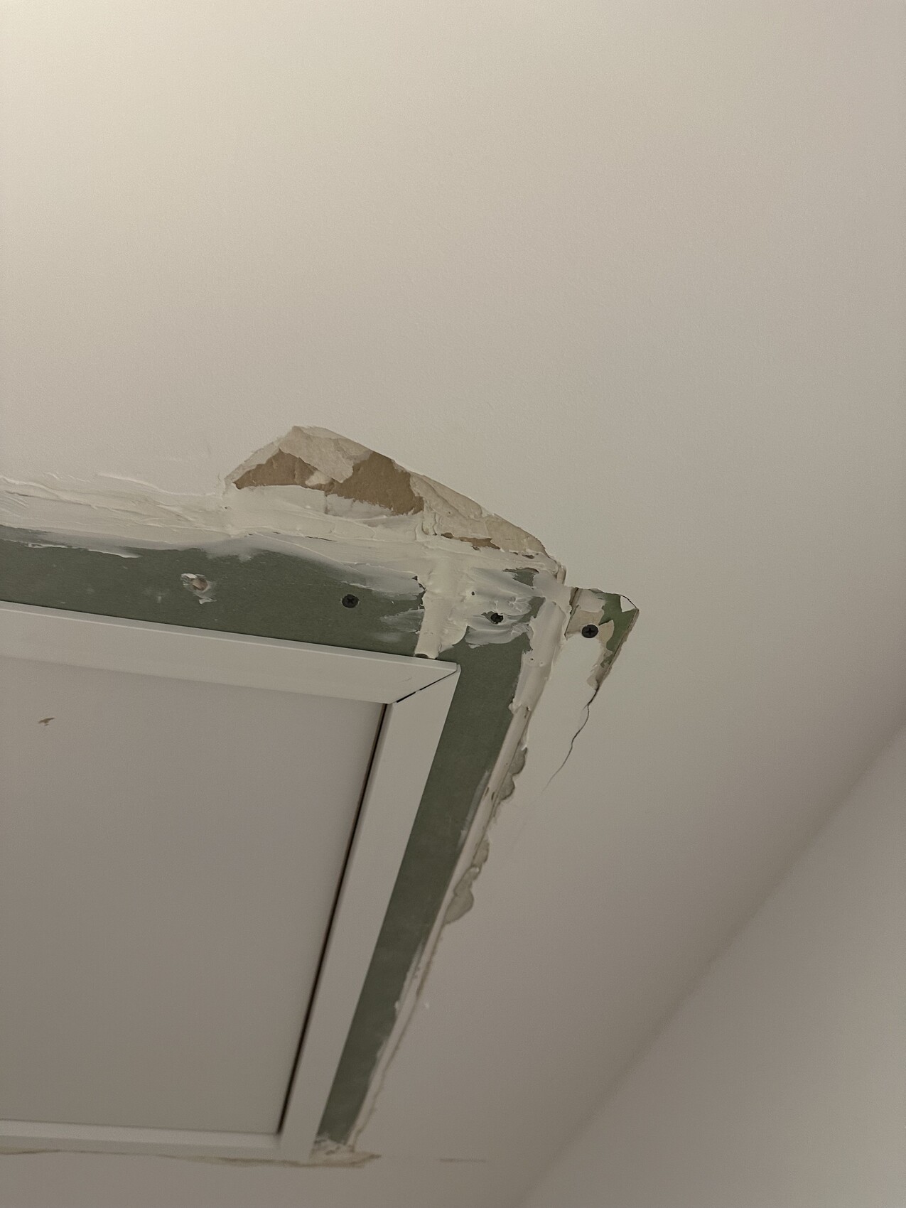 Decke mit beschädigter Kante um eine rechteckige Lüftungsluke; Putz bricht ab.