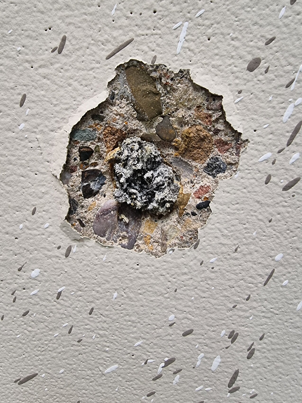 Runde Öffnung in weißer Putzwand; dahinter grobes Kiesel- und Steinmaterial sichtbar.