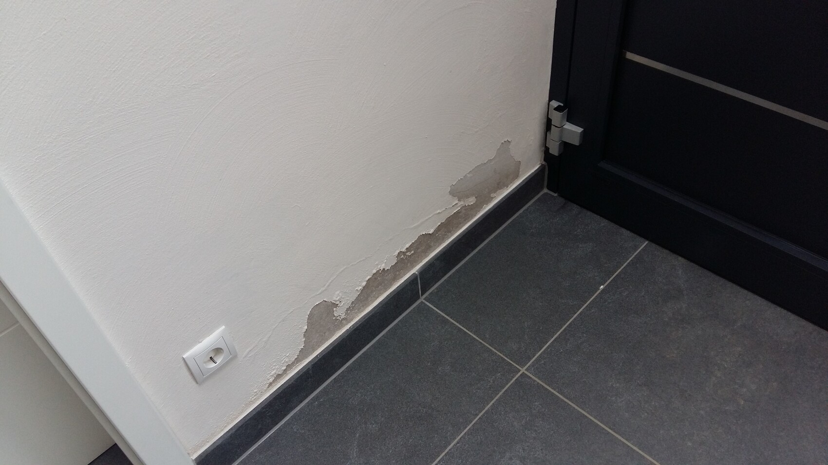 Weiße Wand mit abgeplatztem Putz am unteren Rand neben einer schwarzen Tür; dunkler Fliesenboden.