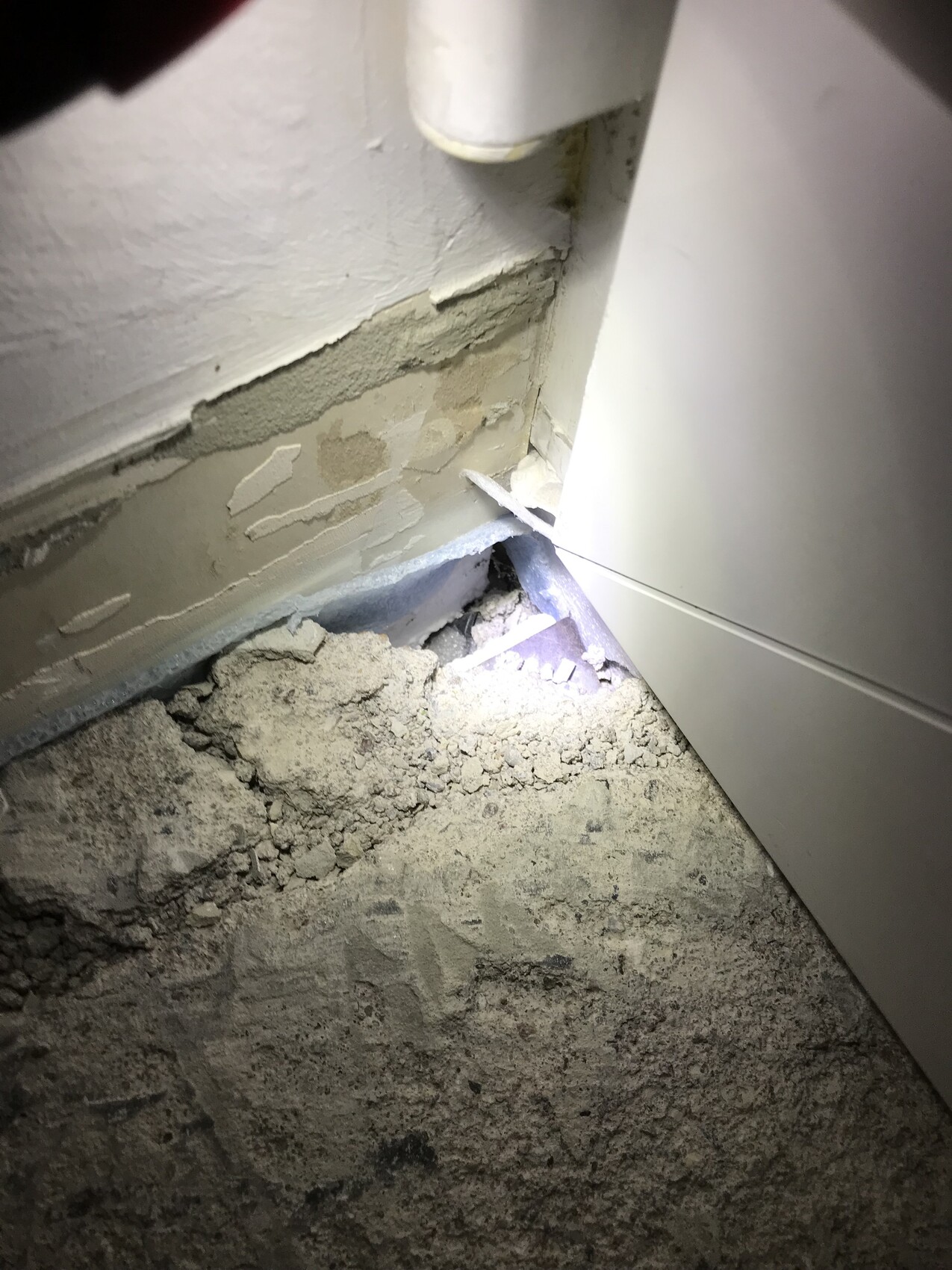 Wand mit abblätterndem Putz, Rohr in Ecke; Staub und Bruchstücke auf dem Boden.