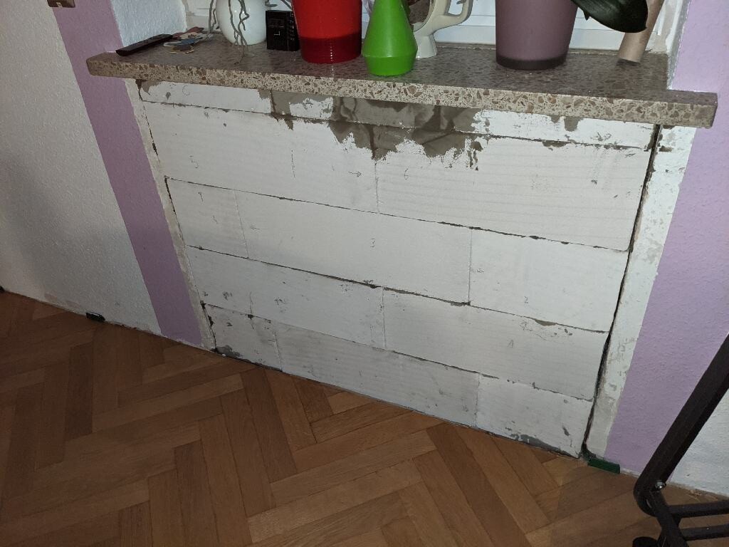 Verputzter Schrank aus weißen Blöcken mit Kratzern; bunte Tassen auf der Platte, Holzboden.