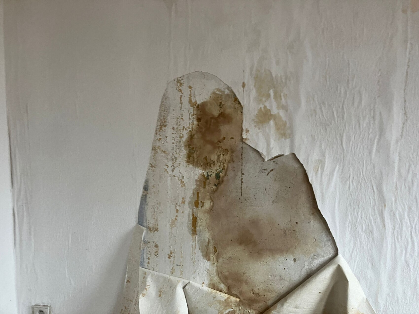 Weiße Wand mit großem abblätternden Putzfleck, braune Verfärbungen; vorn ein geknittertes Tuch.