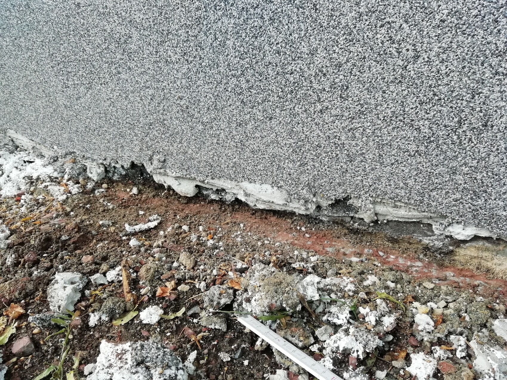 Graue Betonwand mit bröckelnder Kante, Schutt und Messband am Boden.