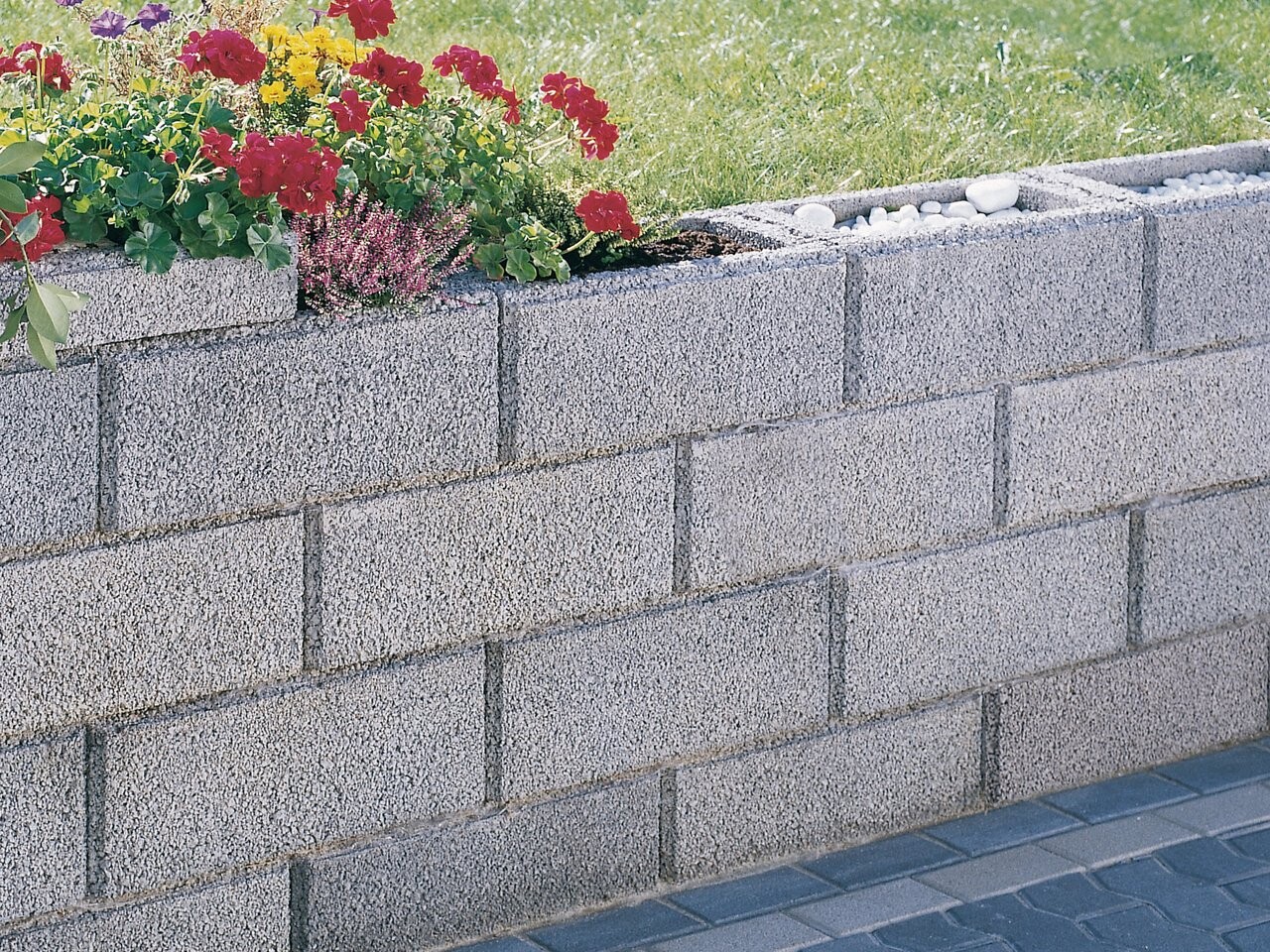 Graue Blockmauer als Beetumrandung mit roten Geranien, grünem Laub und lila Heide, Kies am Rand.