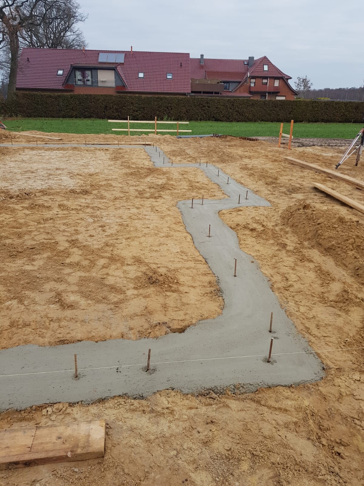 Baustelle: Betonweg im Erdreich gegossen, Bewehrungsstähle ragen, Hintergrund rote Dächer.