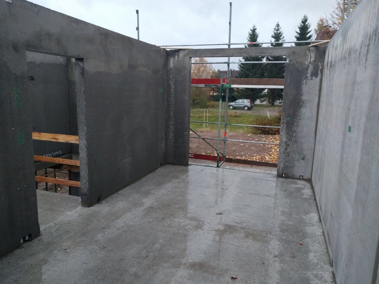 Rohbau aus Beton mit offener Tür zum Außenbereich; Gerüst und Straßenszene im Hintergrund.