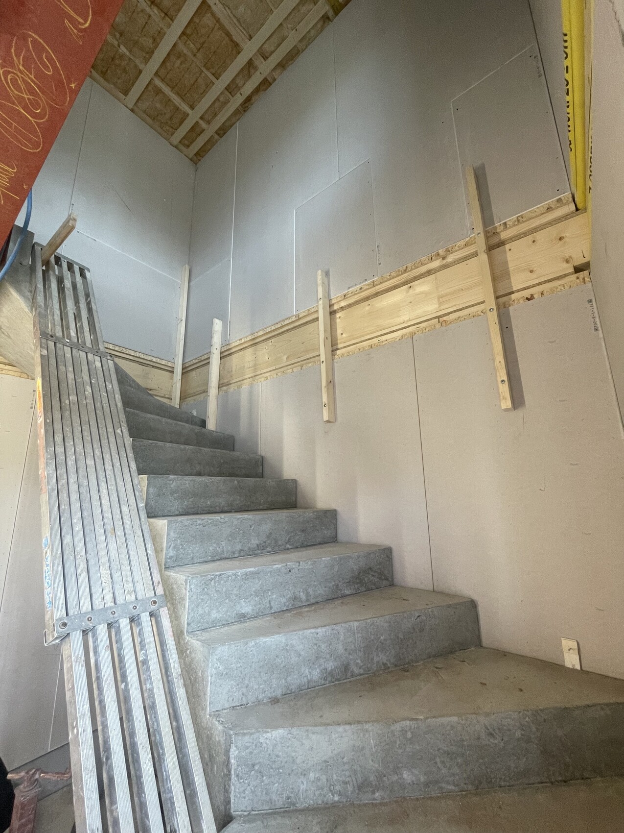 Baustellen-Treppenhaus: Rohbetonstufen, Holzgerüst und Metallleiter an unfertigen Wänden.