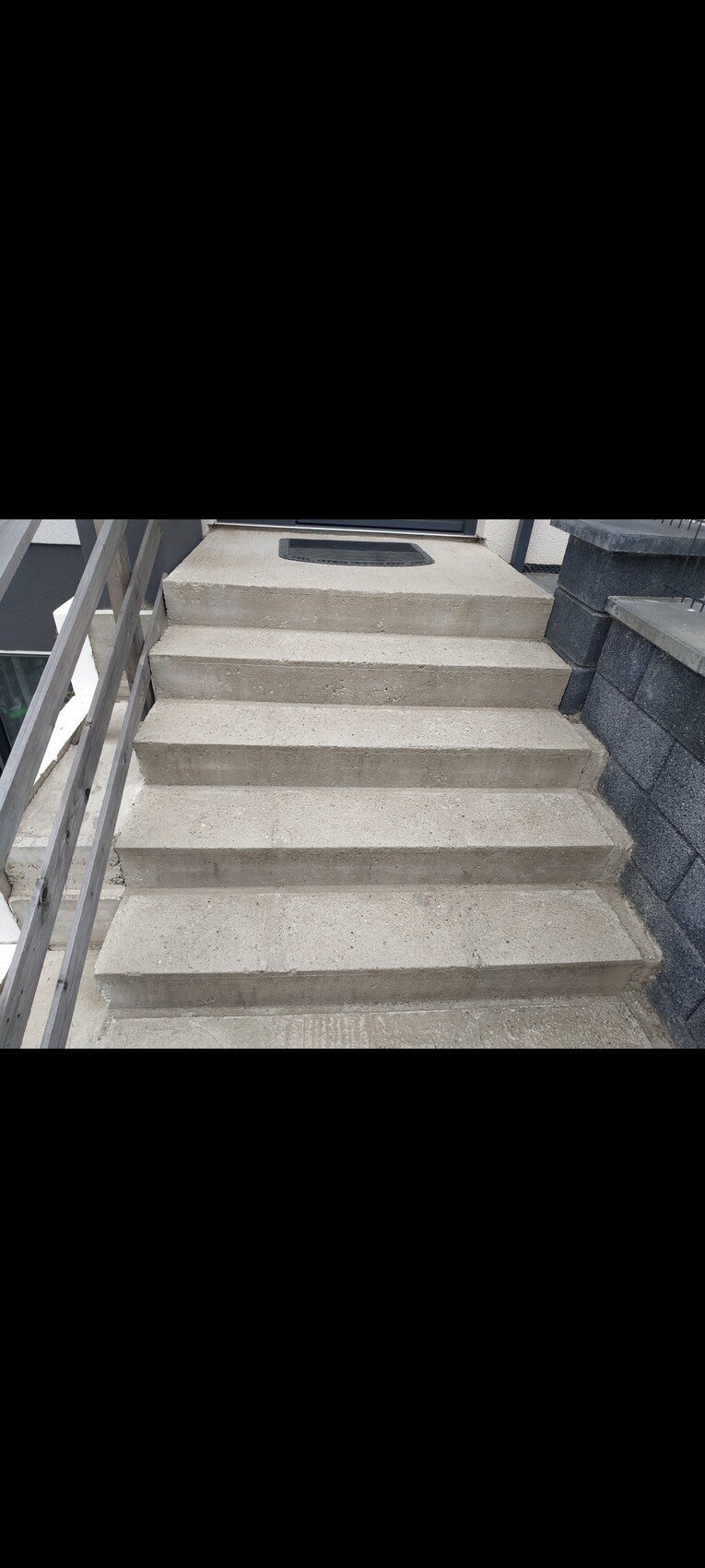 Betonstufen führen zur Haustür; links Holzgeländer, rechts Steinmauer.