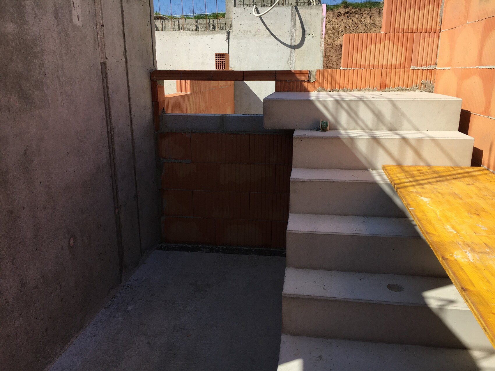Baustelle: Betonwände, weiße Treppen, Holzplatte rechts, orange Ziegelmauer dahinter.