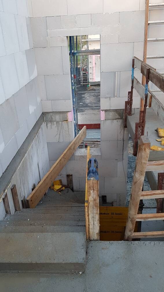 Baustellen-Treppenhaus: Betonstufen, provisorische Holzgeländer, Wandöffnung nach außen.