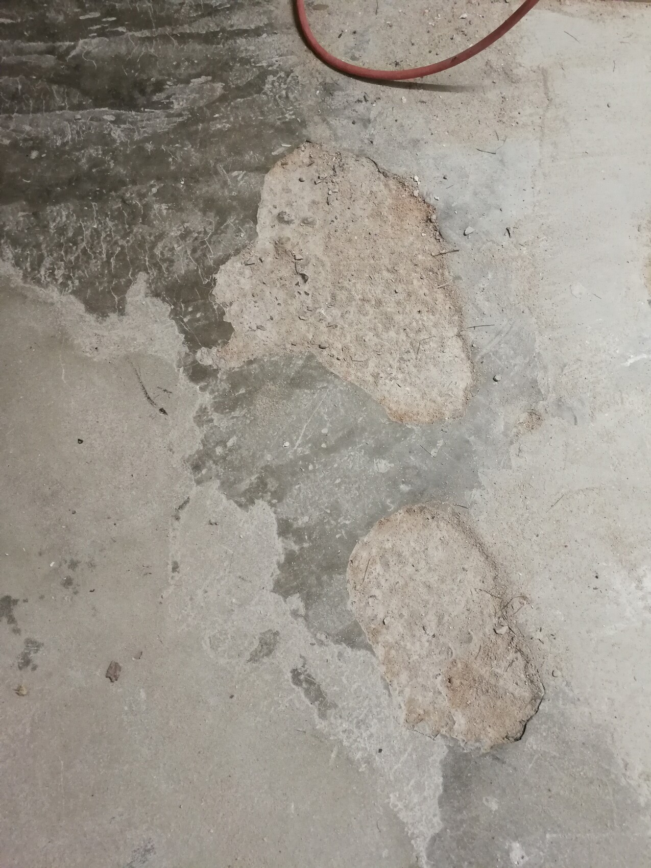 Betonboden mit zwei unregelmäßigen, hellen Putzflecken; roter Schlauch oben.