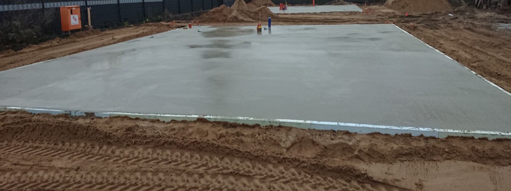 Frisch gegossene Betonplatte auf Baugrube, Randformen sichtbar und Erdarbeiten am Rand.