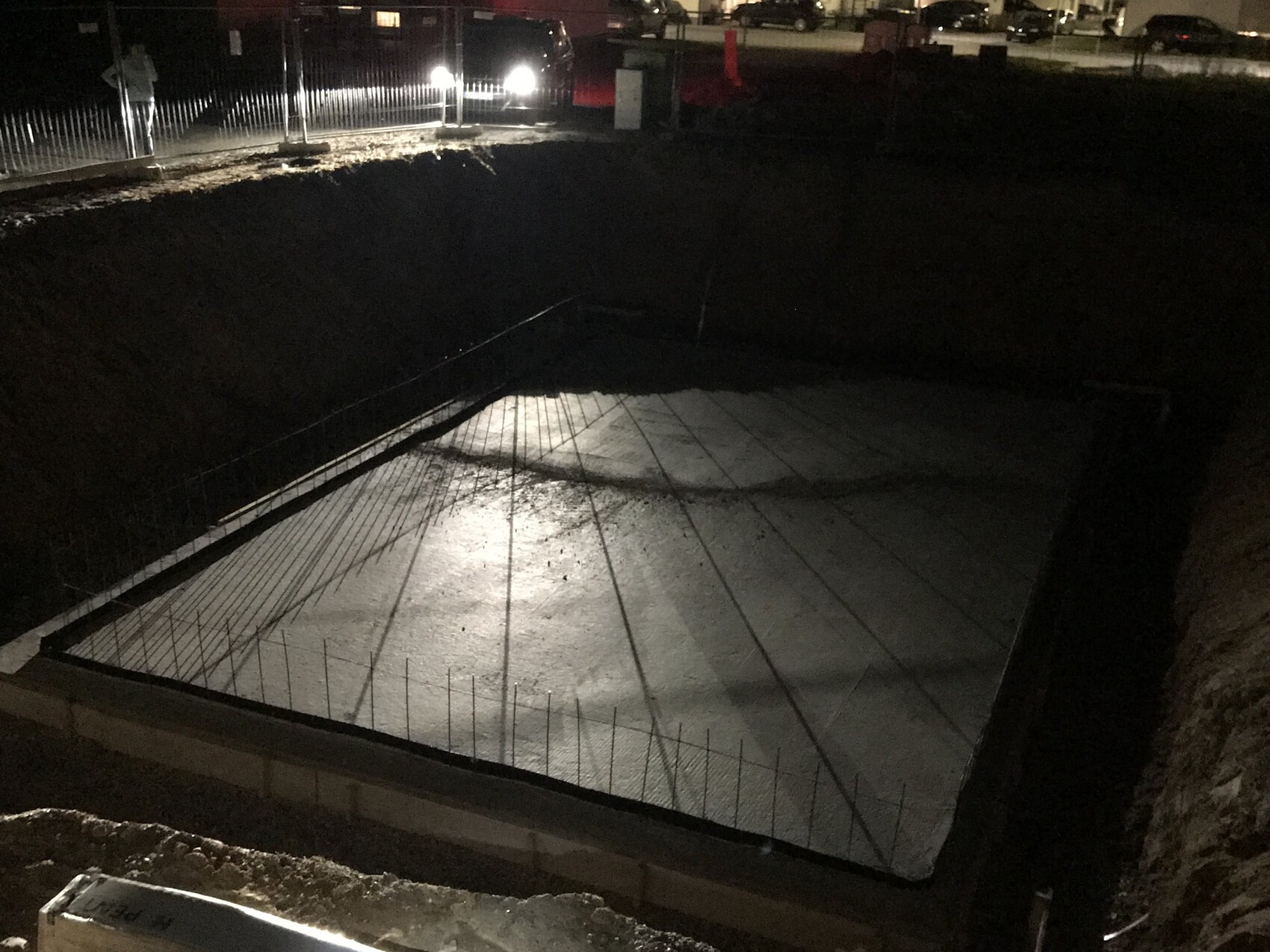Nächtliche Baugrube mit Betonfundament und Bewehrung, Fahrzeuge im Hintergrund.