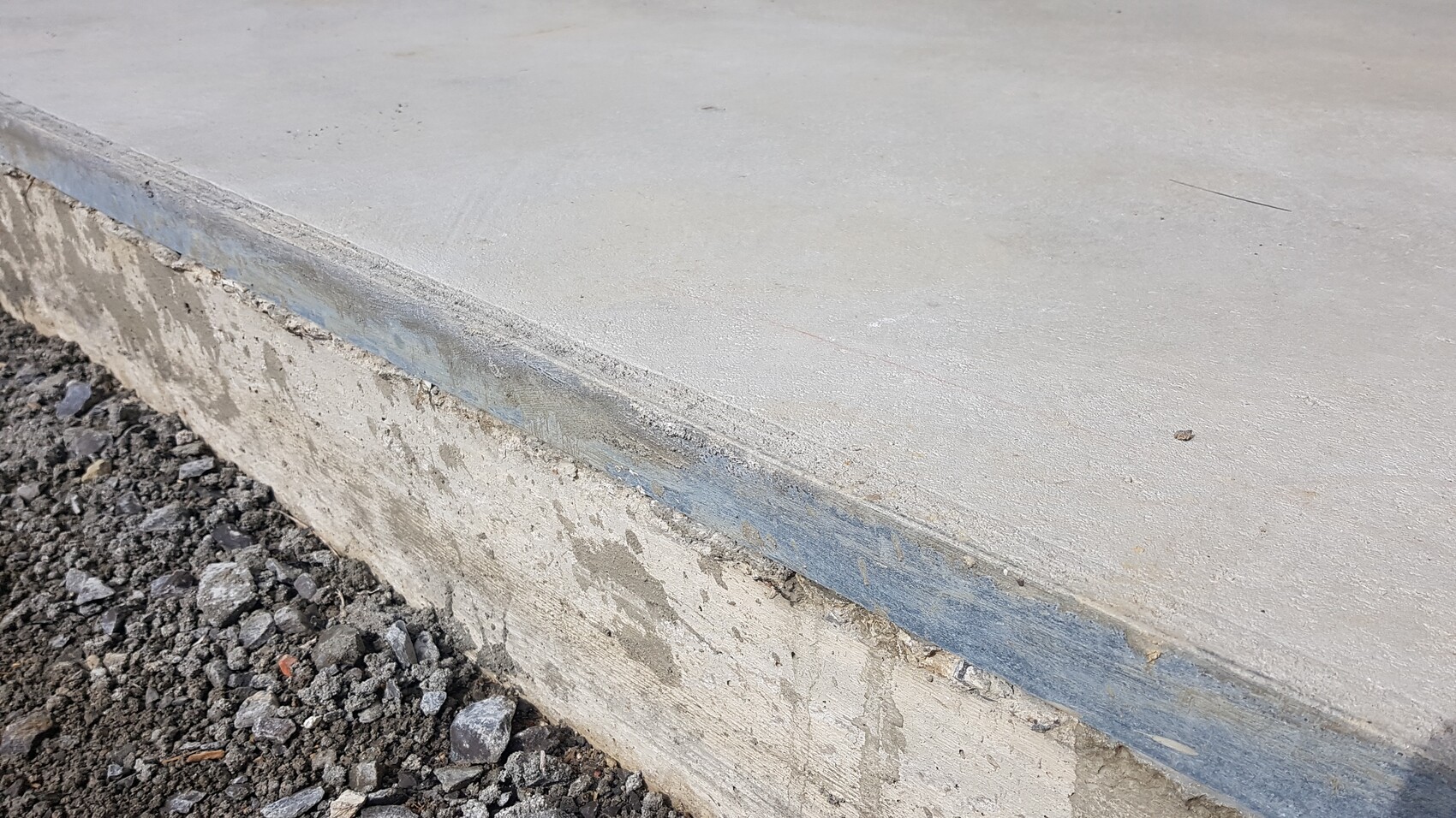 Betonplatte mit Kante, blauem Randstreifen und losem Kies am Boden.